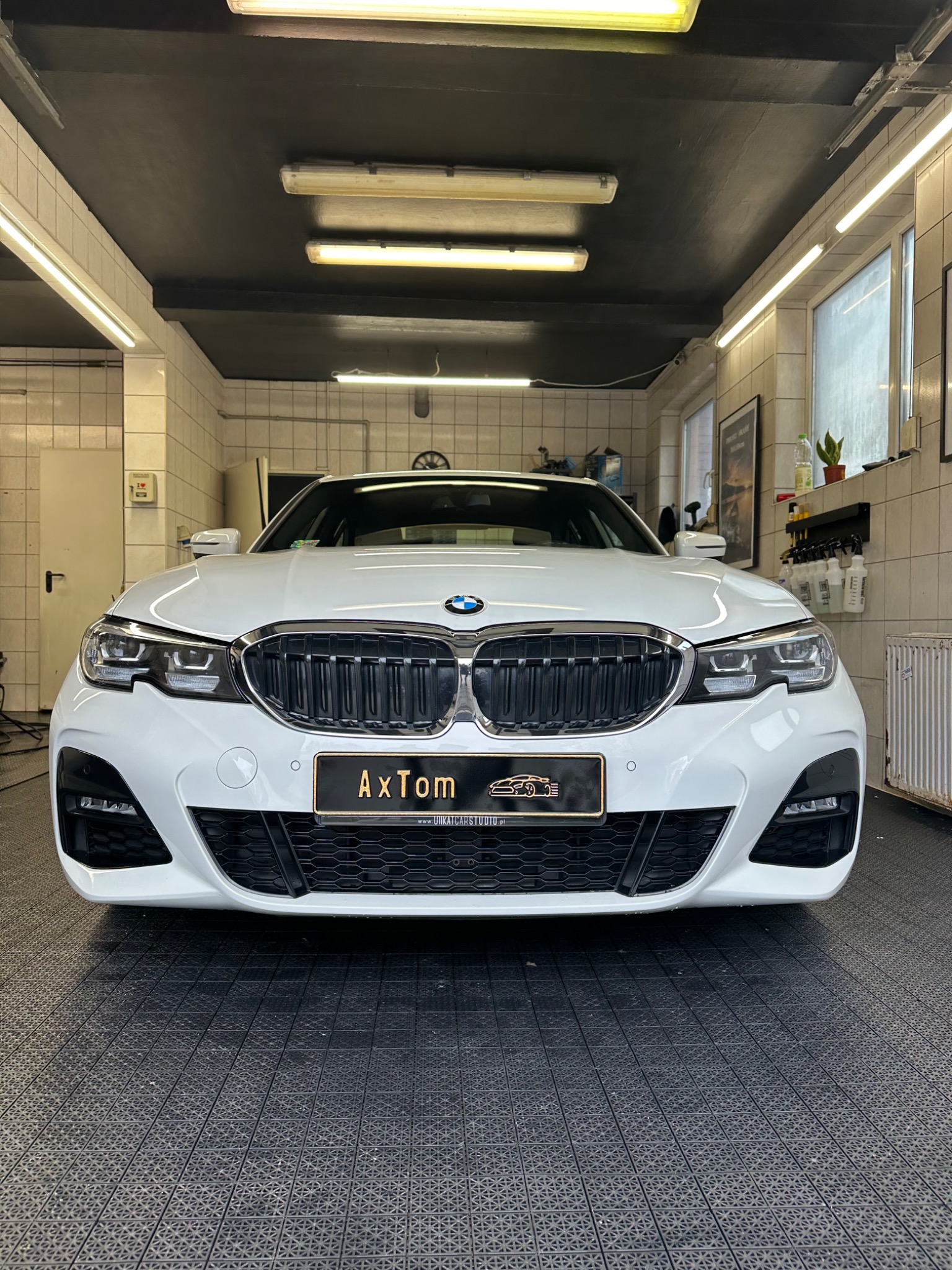 Białe BMW serii 3, widok z przodu, zaparkowane w jasnym, kafelkowanym garażu z czarnym sufitem i oświetleniem LED, na podłodze z czarnej gumowej kostki.