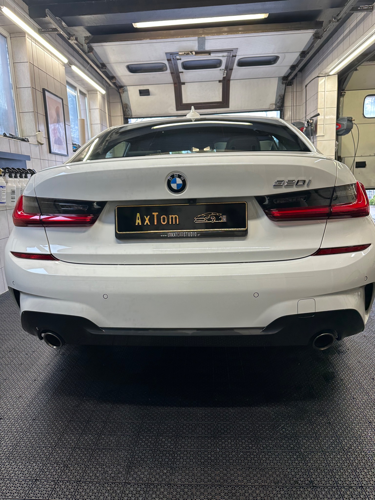 Tył białego BMW 320i w garażu z czarną tablicą rejestracyjną 'AxTom' i logo studia detailingowego.