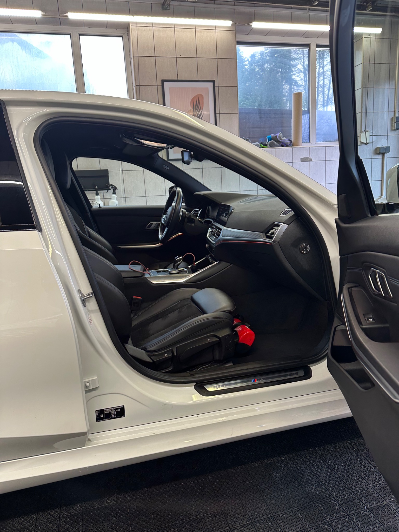 Wnętrze białego samochodu BMW z otwartymi drzwiami, widoczny czarny skórzany fotel, deska rozdzielcza i gaśnica leżąca na podłodze, w tle okna myjni.