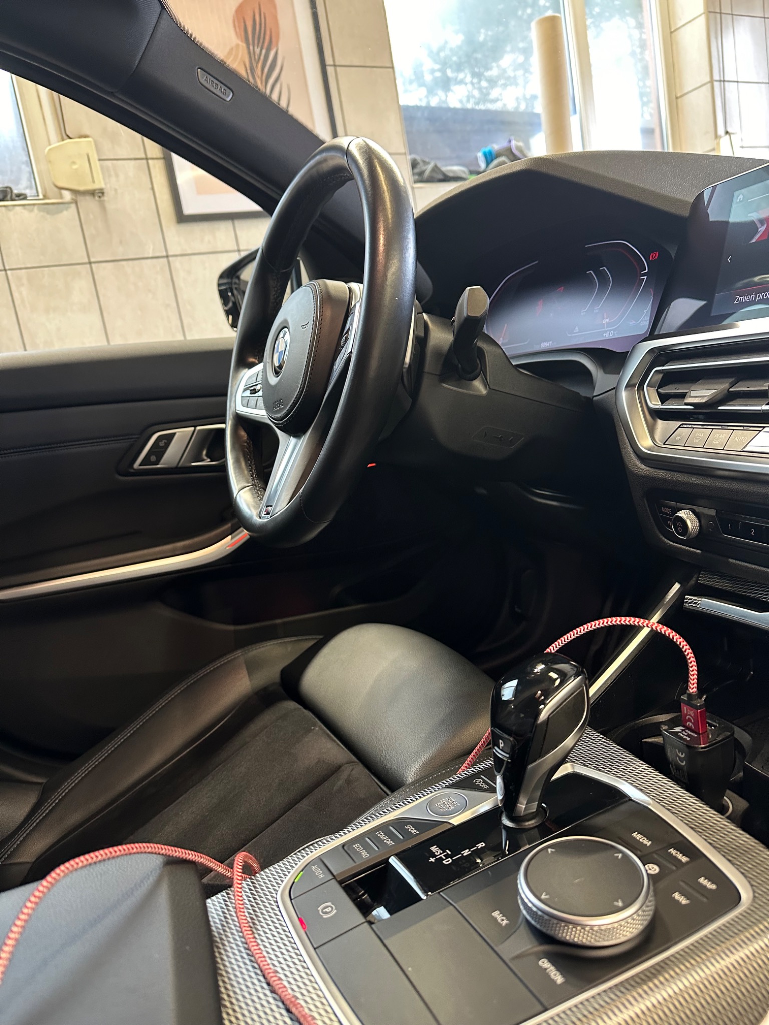 Wnętrze samochodu BMW serii 3 z czarną skórzaną tapicerką, widoczny kokpit, kierownica z logo BMW, konsola środkowa z dźwignią zmiany biegów, ładowarka USB z czerwonym kablem w oplocie...