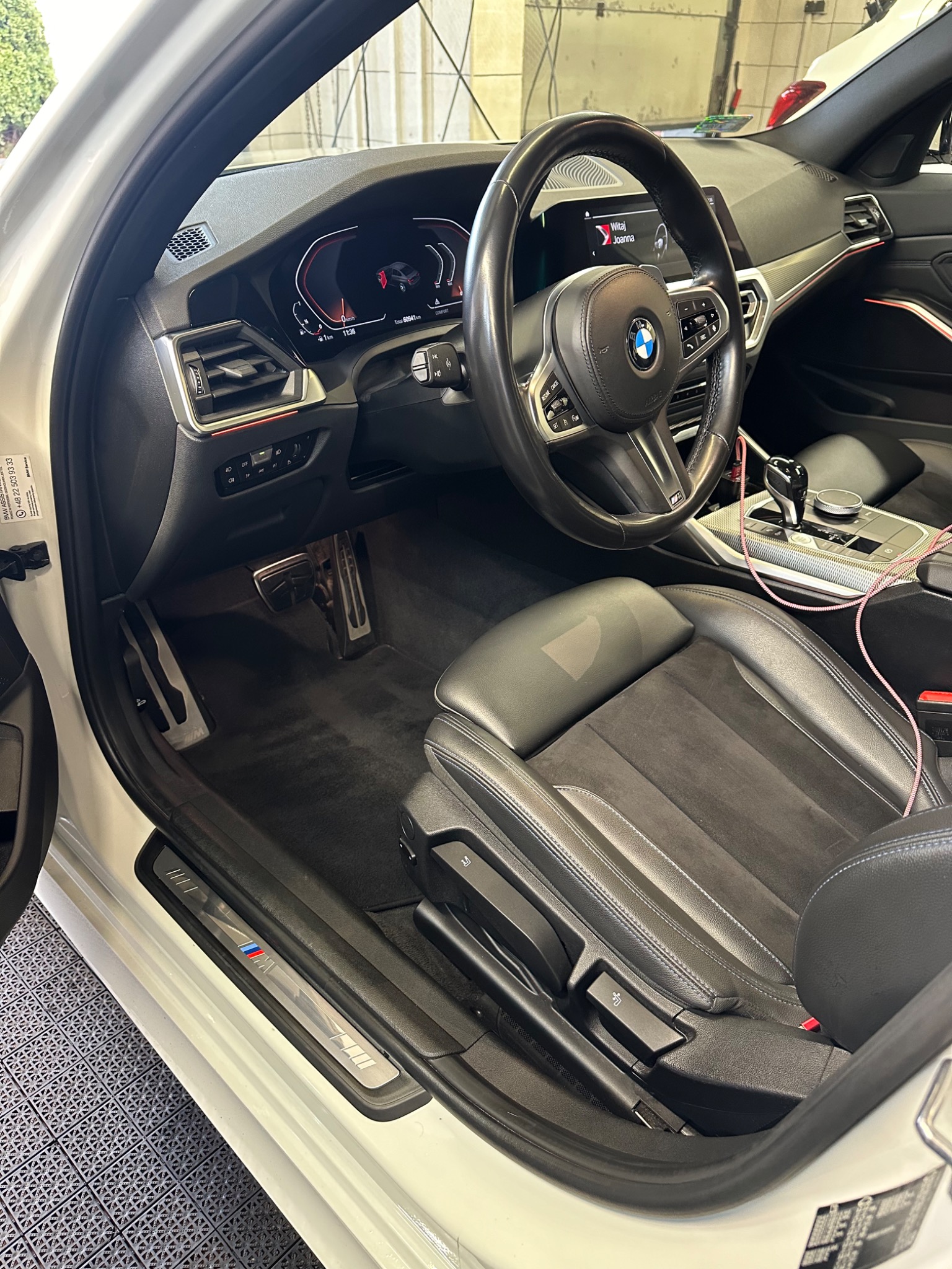 Wnętrze białego samochodu BMW serii 3 z czarną skórzaną tapicerką, widoczny kokpit, fotel kierowcy i fragment progu z logo M Performance. Na fotelu widoczna plama po czyszczeniu.