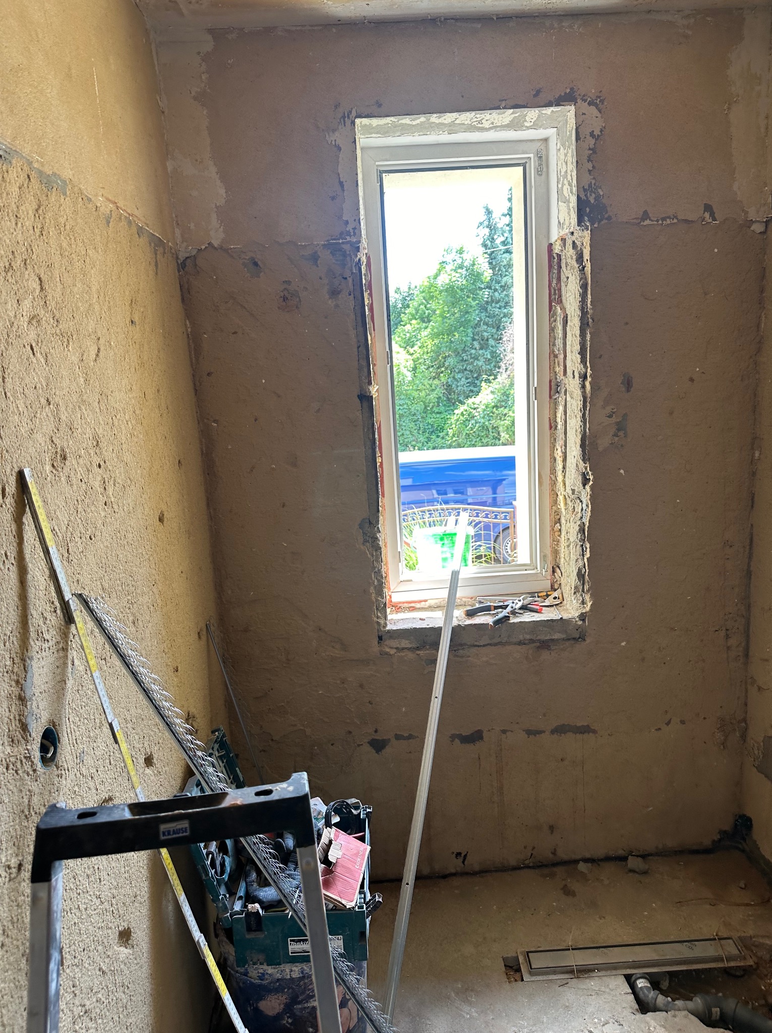 Wnętrze łazienki w trakcie remontu, widoczne surowe ściany z resztkami tynku, nowe okno PCV, drabina Krause oparta o ścianę, skrzynka narzędziowa Makita oraz poziomica.