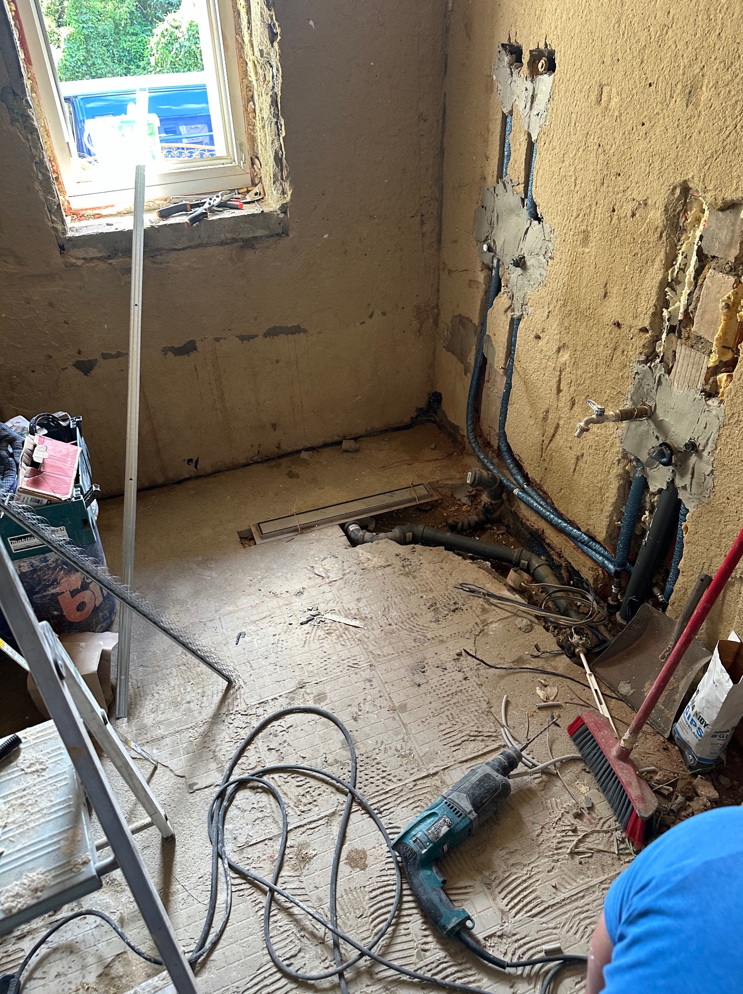 Wnętrze łazienki w Krosno Odrzańskie w trakcie prac instalacyjnych; widoczne nowe rury,  wykute bruzdy w ścianie,  wiertarka,  szczotka i drabina.