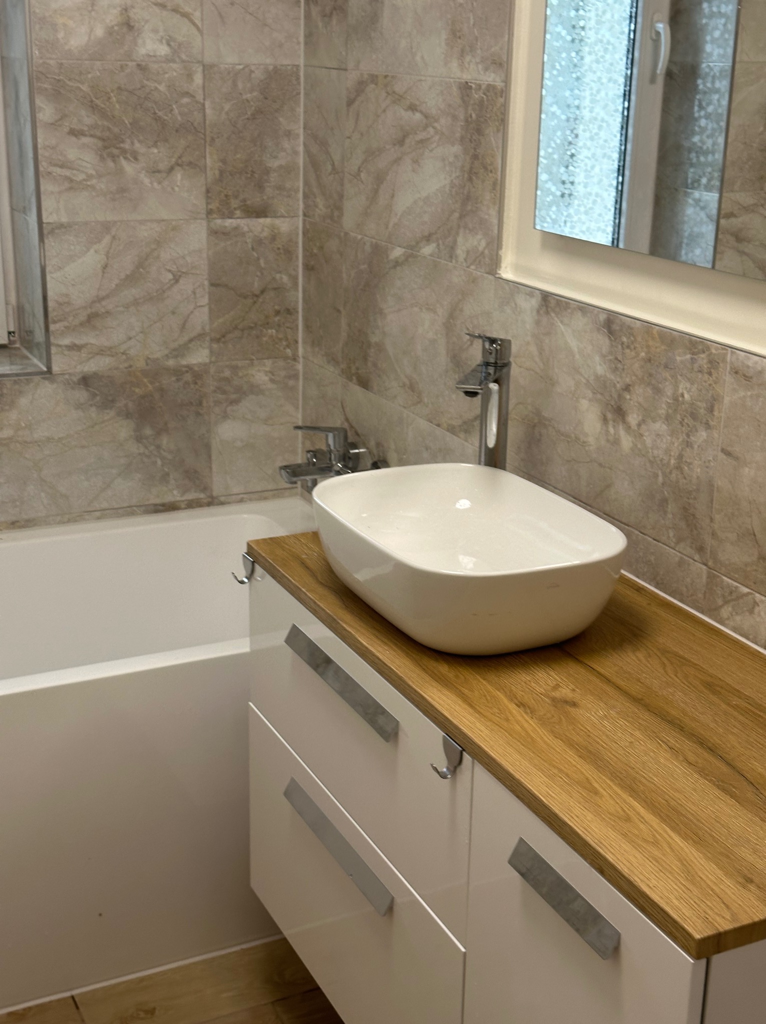 Biała, owalna umywalka nablatowa z chromowaną baterią na drewnianym blacie szafki łazienkowej, na tle marmurowych płytek i wanny.