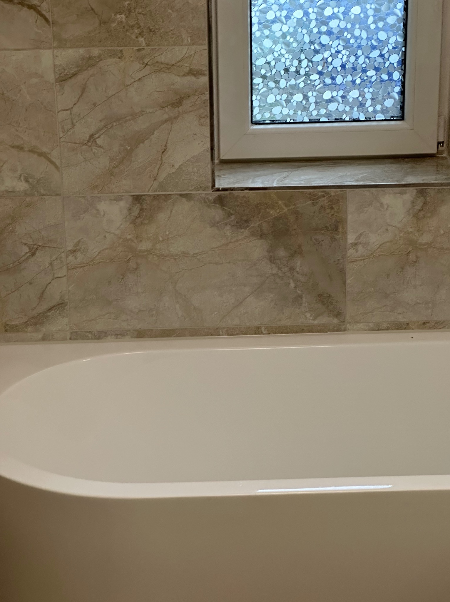 Fragment łazienki z wanną, beżowe płytki imitujące marmur i okno z ozdobną folią.