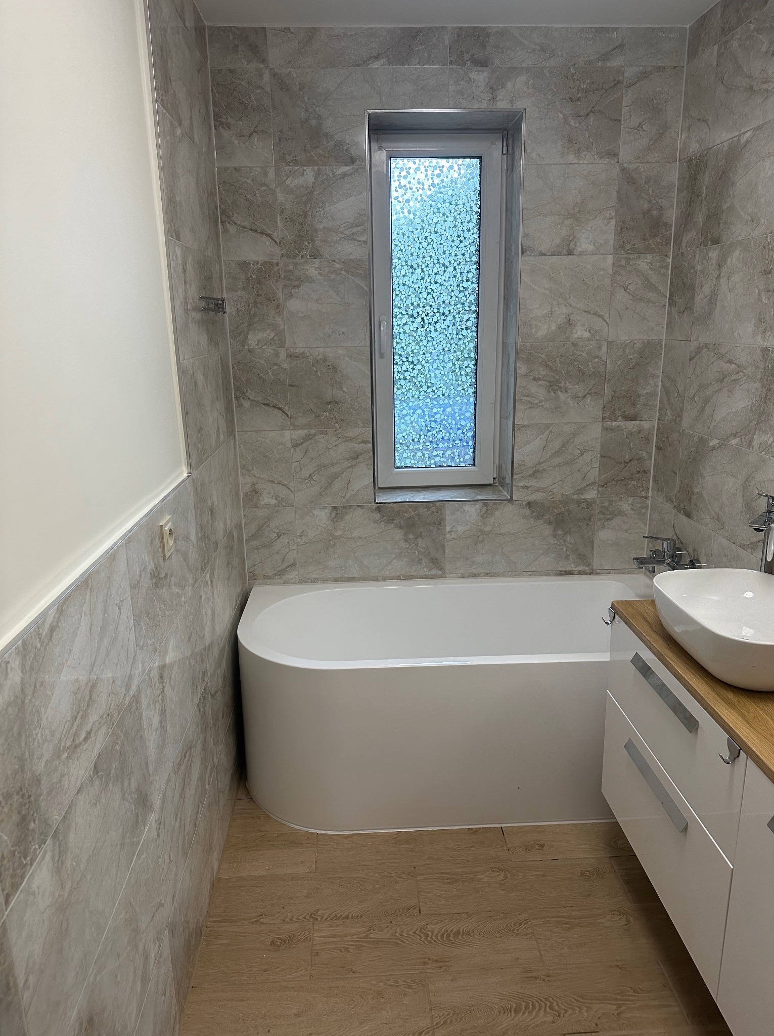 Nowoczesna łazienka z płytkami imitującymi marmur, owalną wanną, umywalką nablatową i podłogą z paneli drewnopodobnych.