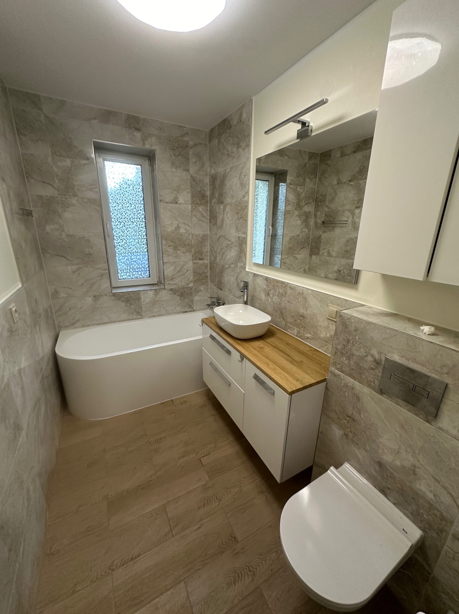 Nowoczesna łazienka w odcieniach beżu i bieli z wanną, umywalką nablatową na szafce z drewnianym blatem oraz wiszącą toaletą.