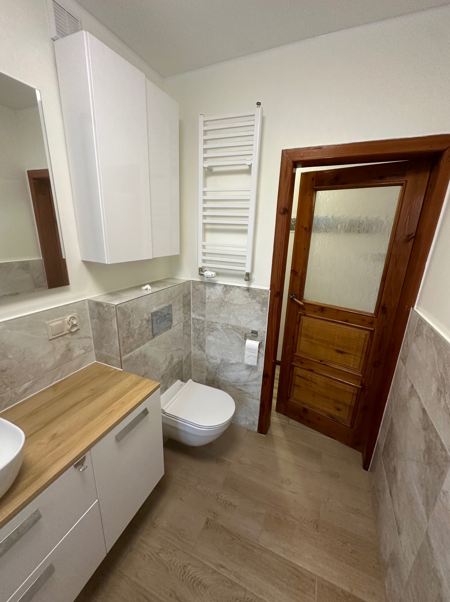 Jasna łazienka po remoncie z drewnianymi akcentami: blat szafki pod umywalką i rama drzwi z szybą z matowego szkła, płytki imitujące marmur na ścianach i podłodze, biała wisząca toaleta i grzejnik...