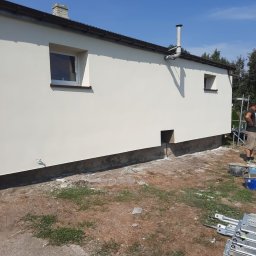 LET MY SALE SPÓŁKA Z OGRANICZONĄ ODPOWIEDZIALNOŚCIĄ - Świeżo otynkowana ściana budynku z dwoma oknami, widoczny fragment rusztowania, wiaderka i elementy rusztowania na ziemi, w tle drzewa i błękitne niebo.