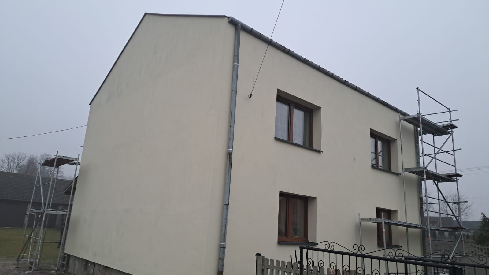 Dwukondygnacyjny dom jednorodzinny z elewacją w kolorze jasnego beżu, widoczne rusztowania przy ścianach, metalowa rynna, ciemna dachówka, pochmurne niebo.