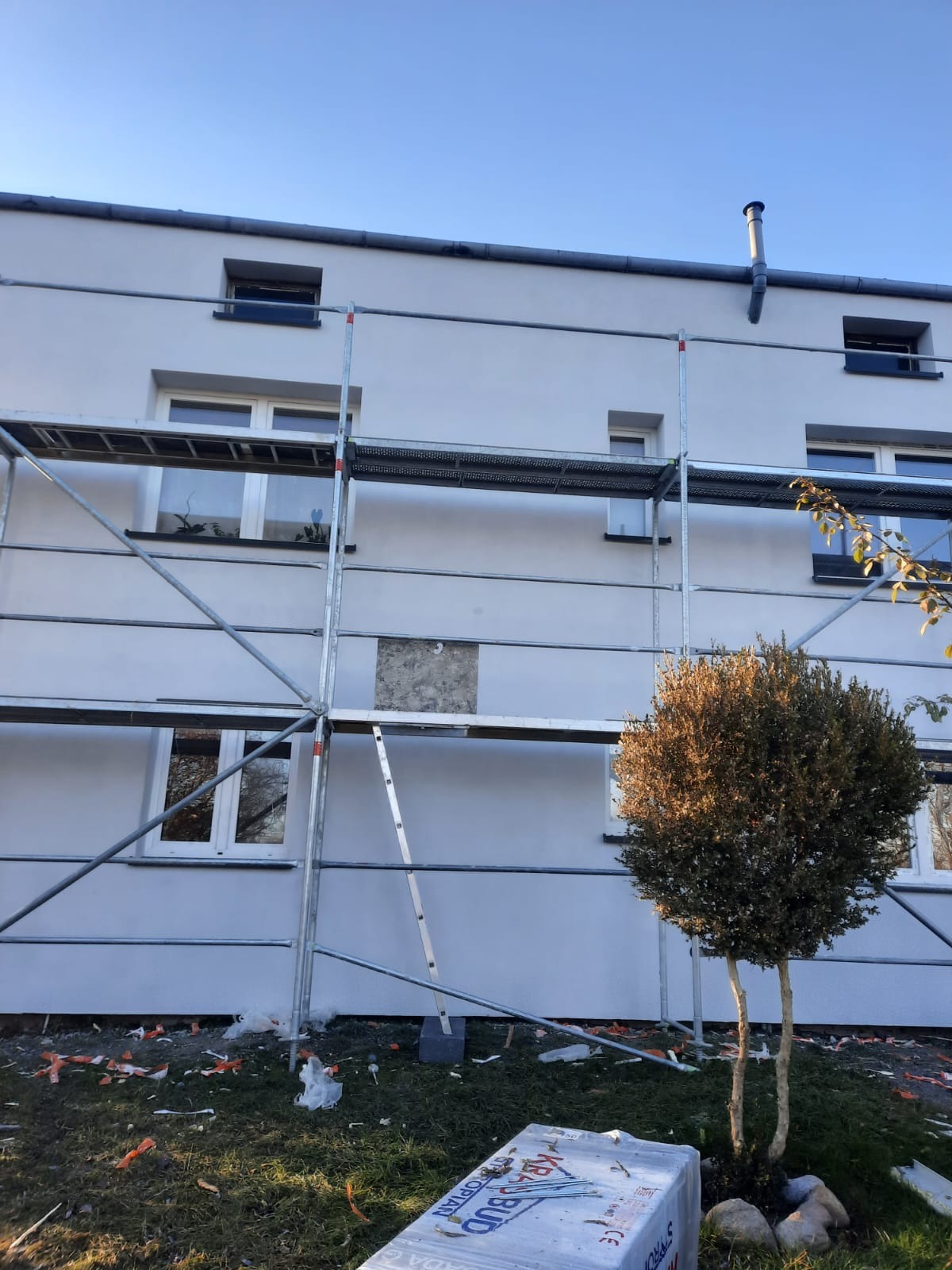 Ocieplanie elewacji budynku z widocznym fragmentem nieocieplonym, rusztowaniem, resztkami materiałów izolacyjnych na trawniku i zapakowanym styropianem u dołu kadru.