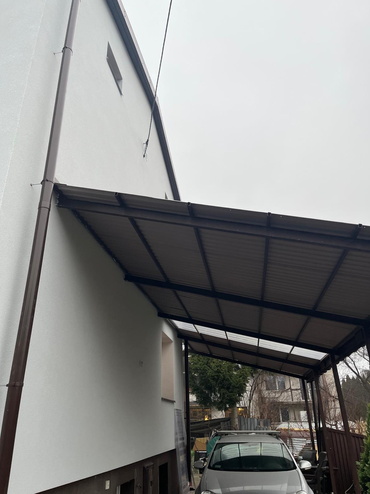 Biała elewacja domu z brązowymi rynnami i zadaszeniem nad miejscem parkingowym; widoczny fragment samochodu, pochmurne niebo.