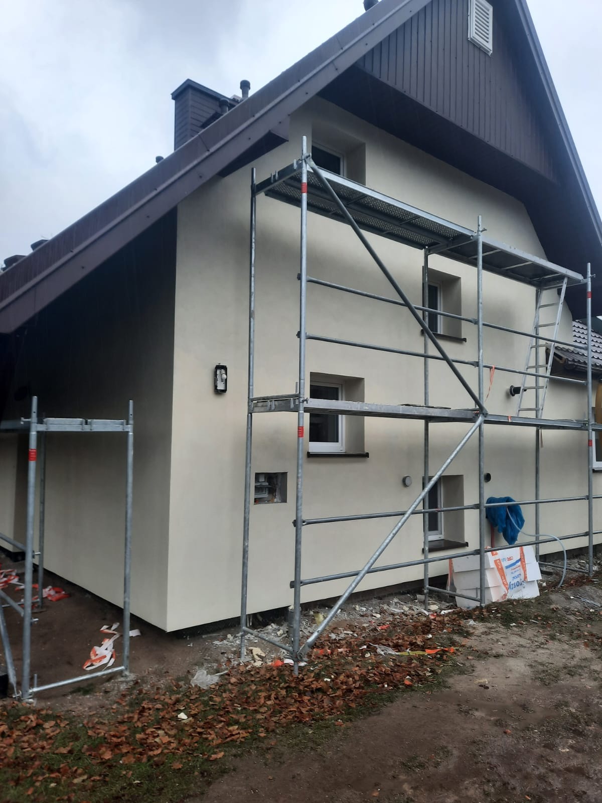 Dom jednorodzinny w trakcie prac elewacyjnych, z widocznymi rusztowaniami i świeżo nałożonym jasnym tynkiem. Na ziemi leżą resztki materiałów budowlanych i opadłe liście.