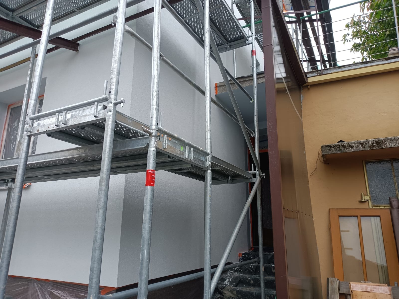 Metalowe rusztowanie przy białej ścianie budynku w trakcie prac, widoczny fragment schodów i sąsiednia, żółta ściana budynku z oknem.