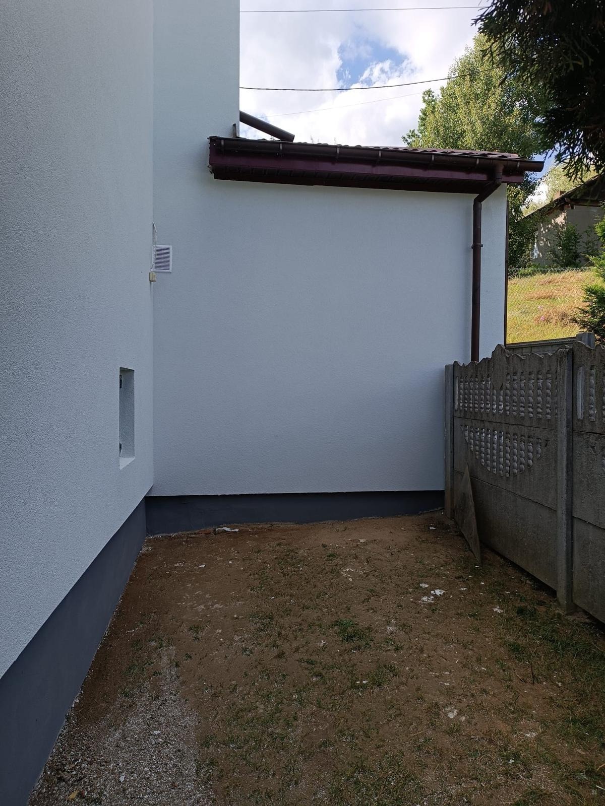 Świeżo otynkowana, jasnoszara ściana budynku z widoczną wentylacją i rynną, przy ziemi ciemnoszara podmurówka, obok betonowe ogrodzenie i ziemia z rzadką trawą.