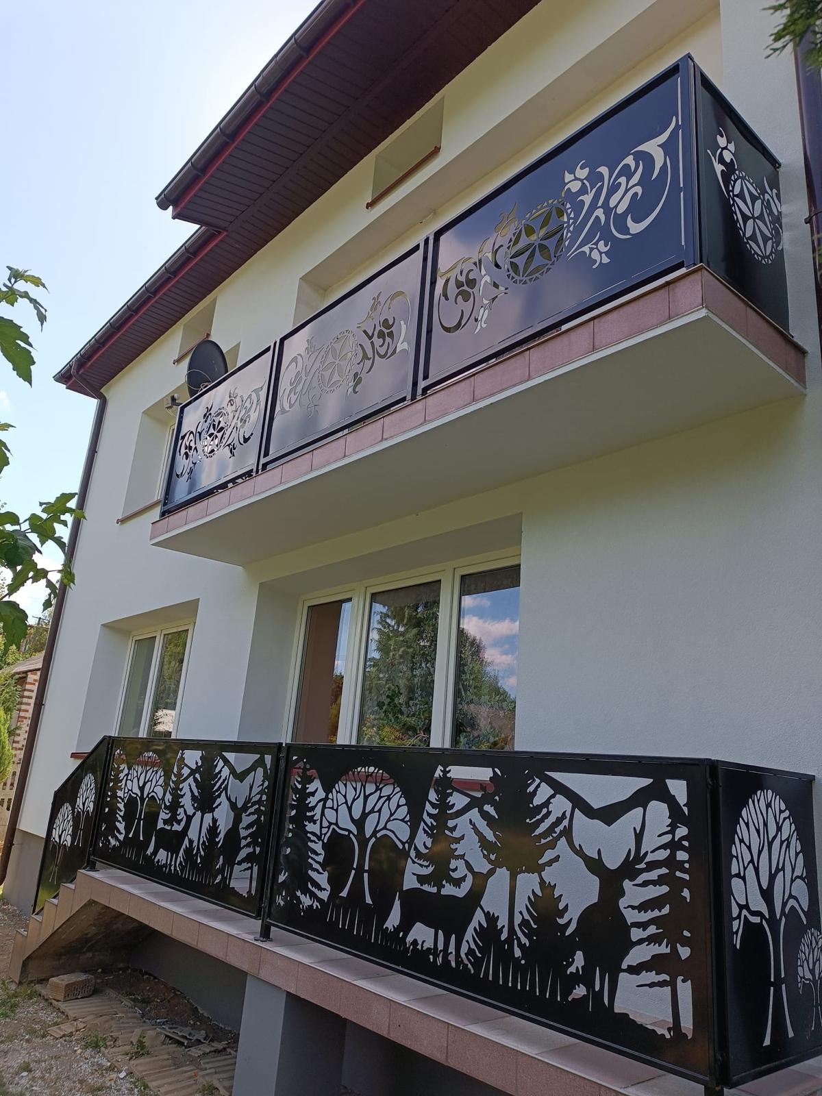 Dwukondygnacyjny dom z białymi ścianami i balkonami z ozdobnymi, metalowymi balustradami. Górny balkon z geometrycznymi i roślinnymi wzorami, dolny z motywem leśnym: jelenie i drzewa.
