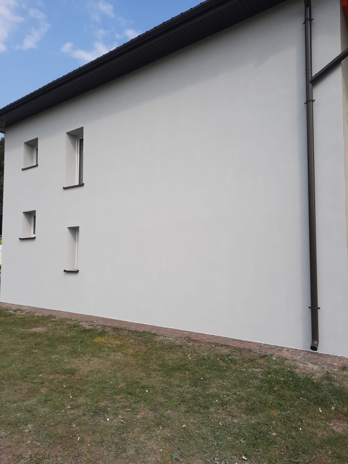 Jasnoszara elewacja budynku z czterema oknami o różnych rozmiarach, brązowe rynny i obróbka dachu, widok z poziomu gruntu na trawnik.