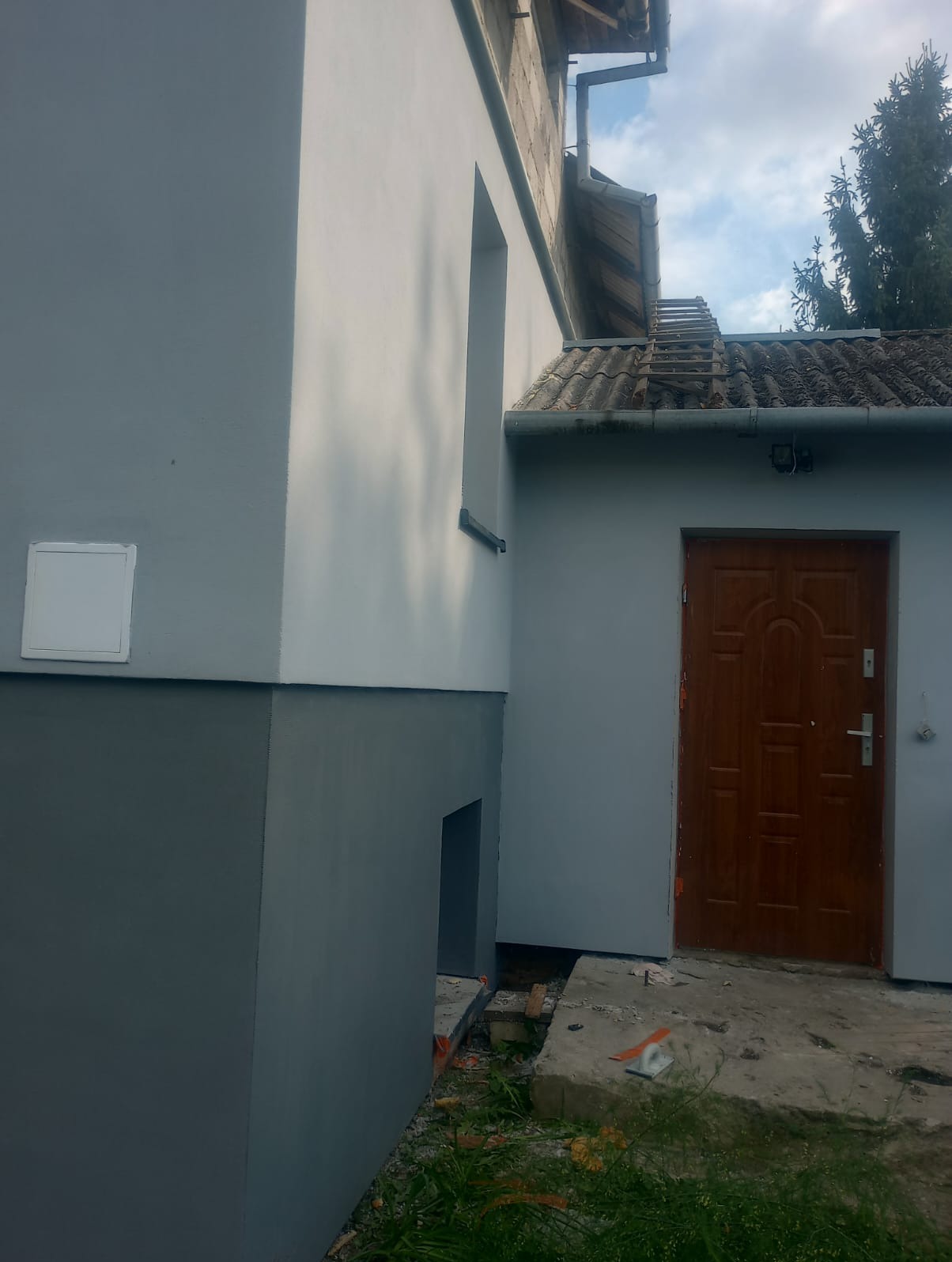 Świeżo pomalowana elewacja budynku z dwukolorowym wykończeniem, szary dół i jaśniejsza góra, widoczny fragment dachu z dachówką i drewniane drzwi wejściowe.
