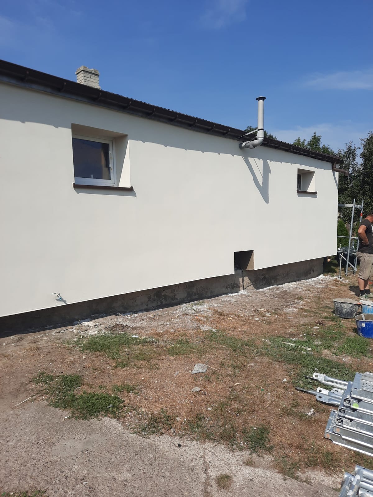 Świeżo otynkowana ściana budynku z dwoma oknami, widoczny fragment rusztowania, wiaderka i elementy rusztowania na ziemi, w tle drzewa i błękitne niebo.
