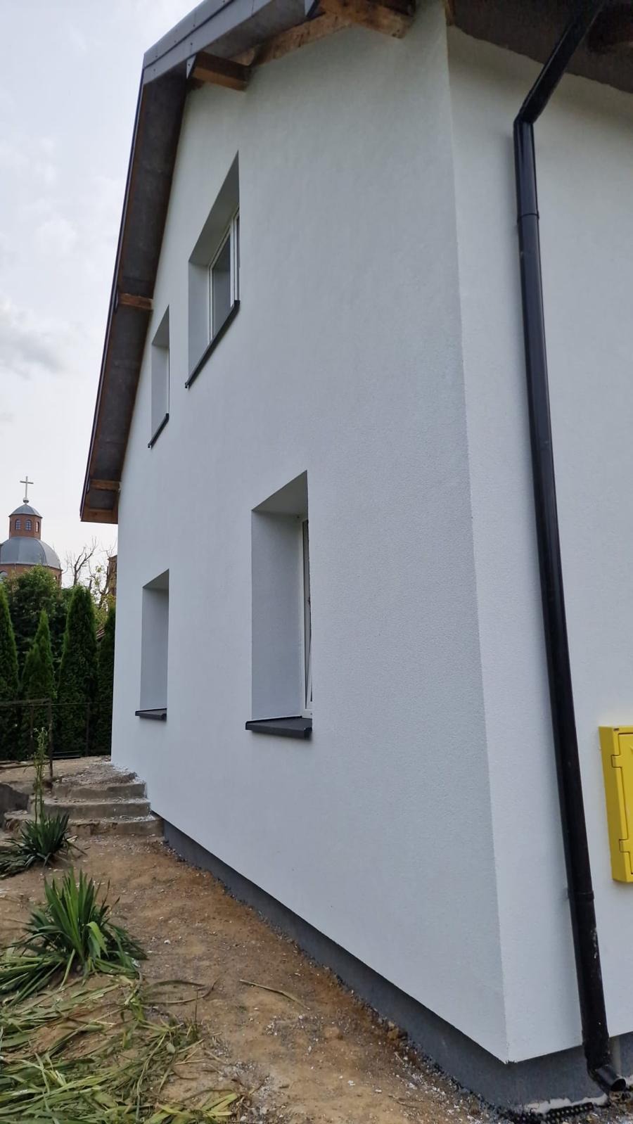 Fragment bocznej ściany domu po nałożeniu jasnoszarego tynku strukturalnego, widoczne okna z ciemnymi parapetami, czarna rynna, żółta skrzynka gazowa i fragment dachu, w tle kopuła kościoła i zieleń.