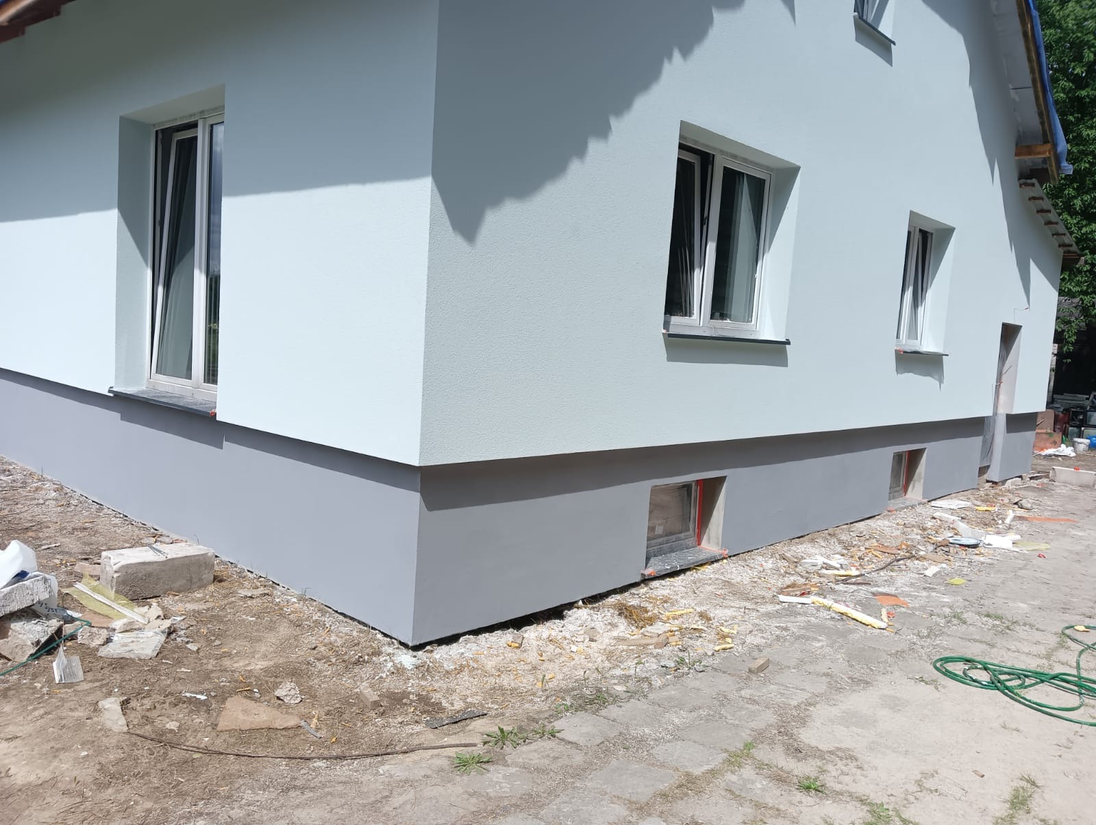 Świeżo otynkowany i pomalowany fragment domu jednorodzinnego z widocznym szarym cokołem, jasnoszarą elewacją i białymi oknami, teren wokół domu częściowo zagospodarowany, widoczne resztki...