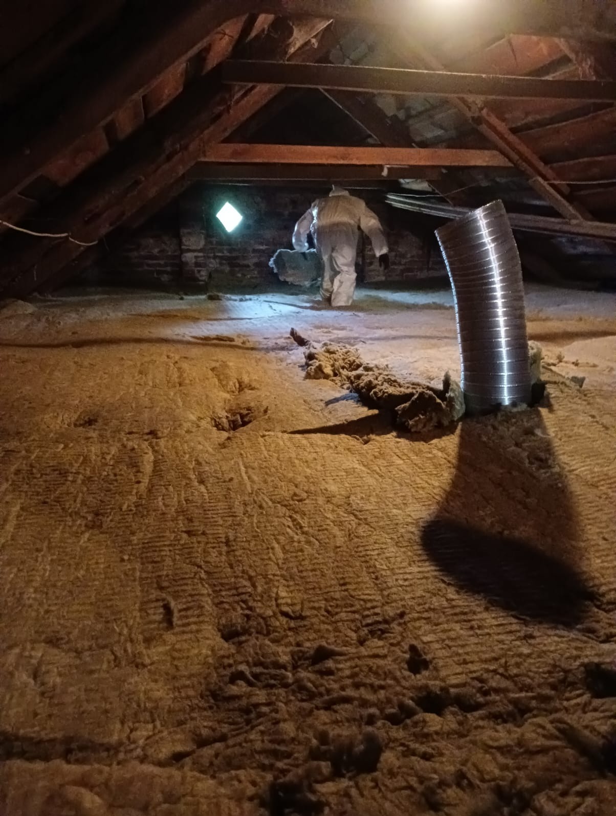 Pracownik w kombinezonie ochronnym instaluje izolację z wełny mineralnej na poddaszu, widoczny fragment okna i przewód wentylacyjny.