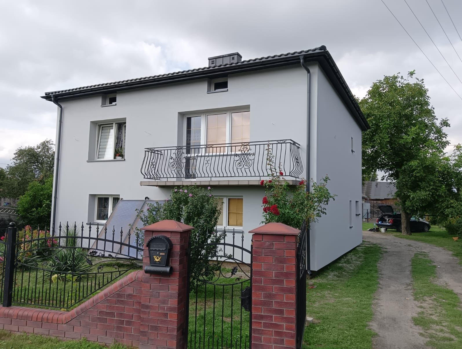 Dwupiętrowy dom z balkonem i szarą elewacją, widoczny zza ogrodzenia z ceglanymi słupkami i czarną skrzynką pocztową. Na elewacji widoczne panele słoneczne.