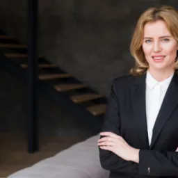 Portret kobiety w czarnym garniturze, stojącej na tle loftowego wnętrza z metalowymi schodami i betonową ścianą.