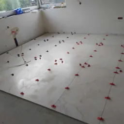 Białe płytki podłogowe imitujące marmur, ułożone w trakcie prac, z użyciem czerwonych i niebieskich klipsów poziomujących oraz czarnych sznurków wyznaczających linie, widoczne instalacje na ścianie...