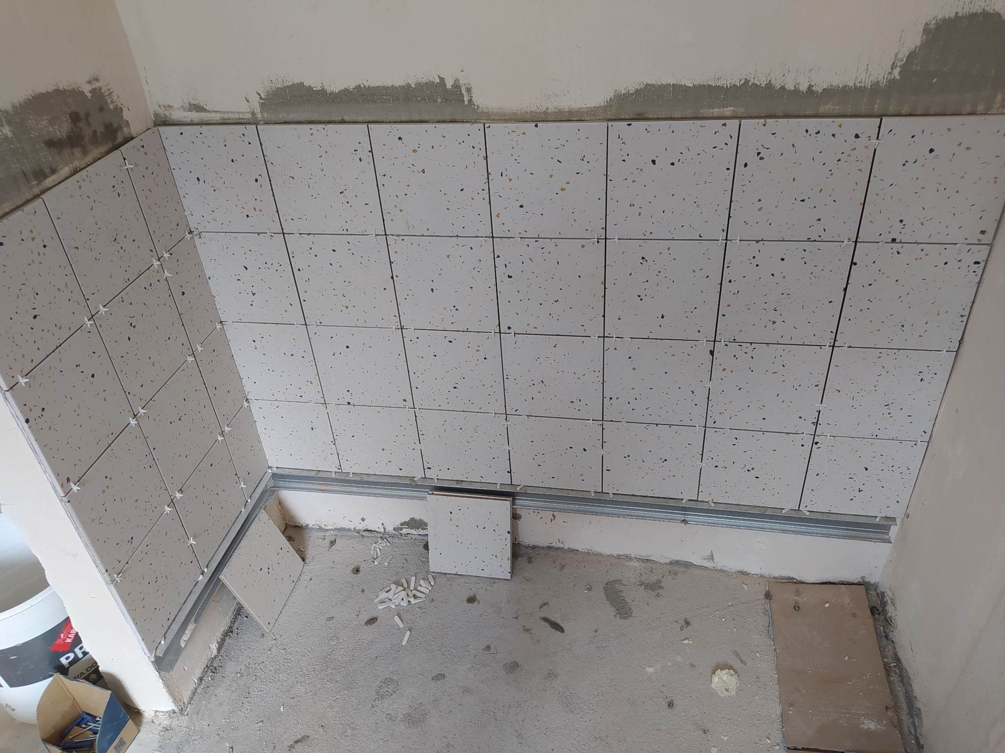 Ściana łazienki w trakcie układania płytek terazzo z użyciem krzyżyków dystansowych i metalowej listwy przypodłogowej, widoczny fragment surowego stanu ściany i podłogi.