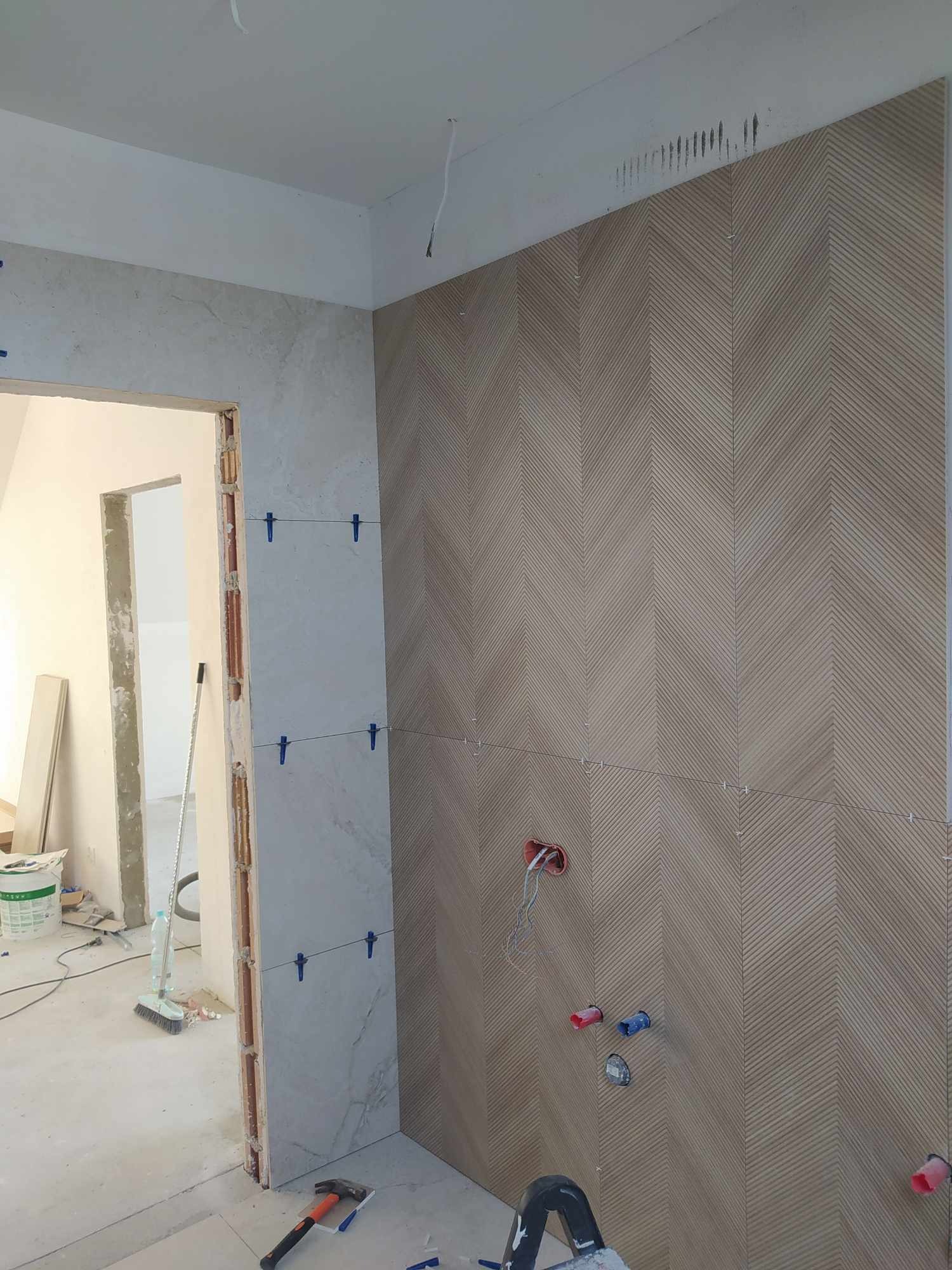 Ściana łazienki wyłożona płytkami w jodełkę podczas montażu, widoczne niebieskie krzyżyki dystansowe, wystające kable i instalacje wodne, w tle fragment pomieszczenia w trakcie prac wykończeniowych.