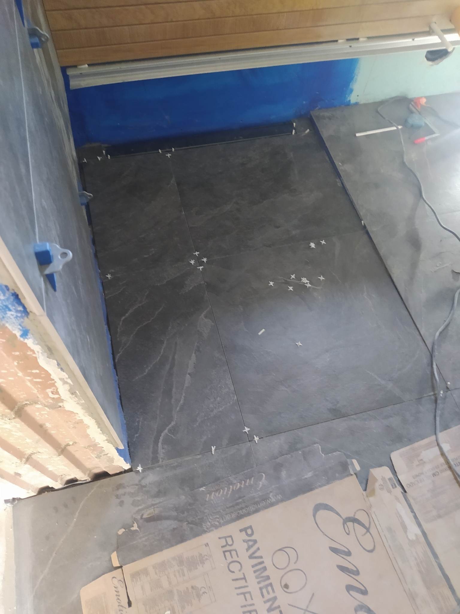 Układanie ciemnoszarych płytek podłogowych w trakcie prac, widoczne krzyżyki dystansowe i opakowanie płytek Emotivo Pavimento Rectified 60x60.