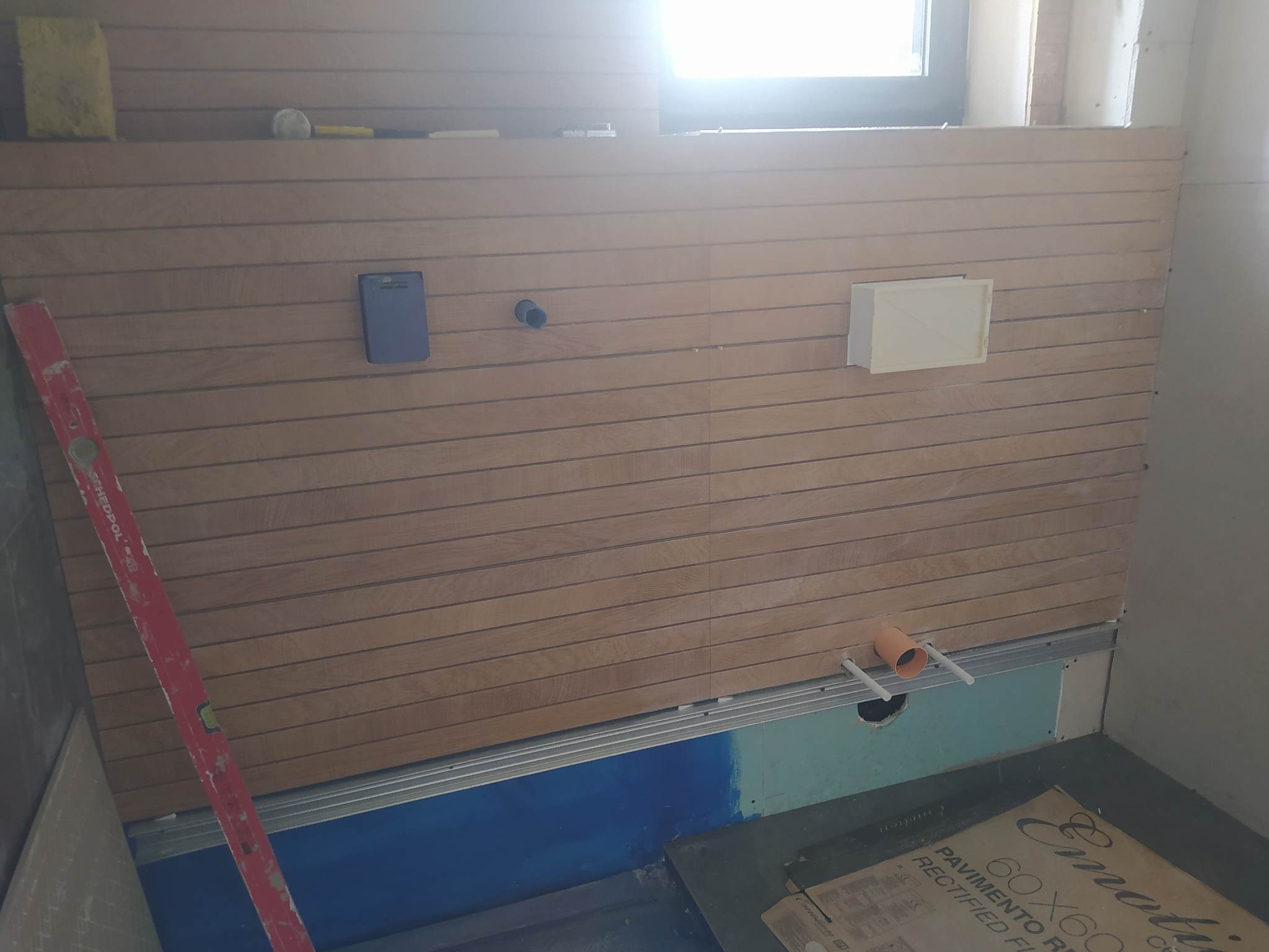 Ściana z drewnianych paneli ułożonych poziomo w trakcie montażu w pomieszczeniu, widoczny poziomica, puszka elektryczna i otwór na rurę kanalizacyjną, pod spodem niebieska izolacja przeciwwilgociowa.