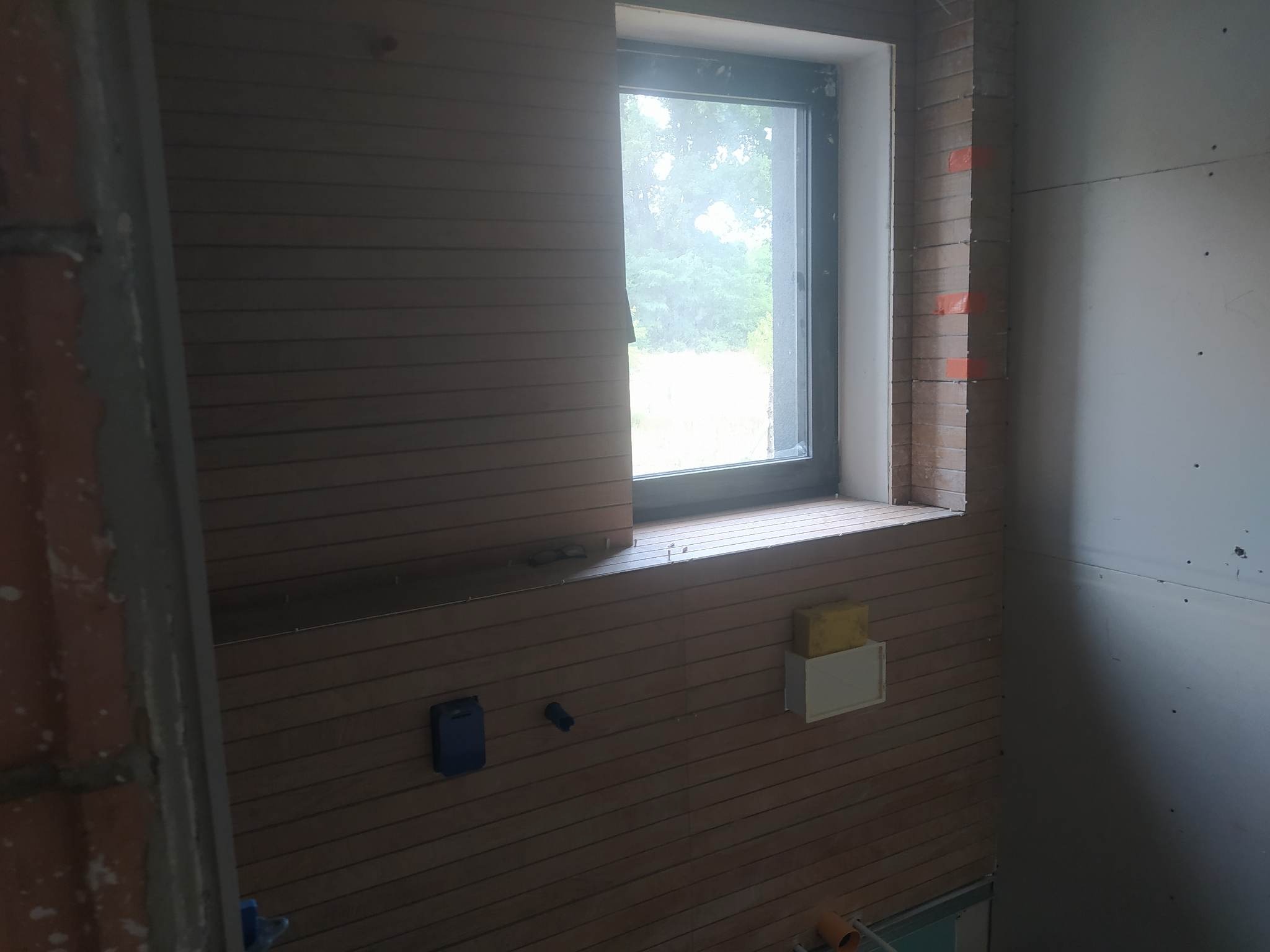 Wnętrze w trakcie prac wykończeniowych: ściana z paneli drewnopodobnych, okno z widokiem na zieleń, ściana z płyt gipsowo-kartonowych z widocznymi łączeniami i fragmentami cegieł.