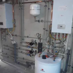Złożona instalacja grzewcza z dwoma piecami, zbiornikiem buforowym, naczyniem przeponowym i licznymi rurami miedzianymi i plastikowymi na surowej ścianie. Widoczne zawory, manometry i elementy...