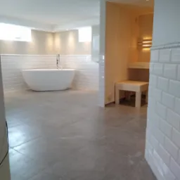 Nowoczesna łazienka z wolnostojącą wanną, wykończona białymi, prostokątnymi płytkami i szarą podłogą, z widoczną sauną z jasnego drewna w głębi.