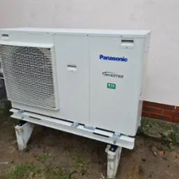 Zewnętrzna jednostka klimatyzacji Panasonic Inverter R32 zamontowana na betonowych podstawach przy ścianie budynku, widoczne ślady deszczu.