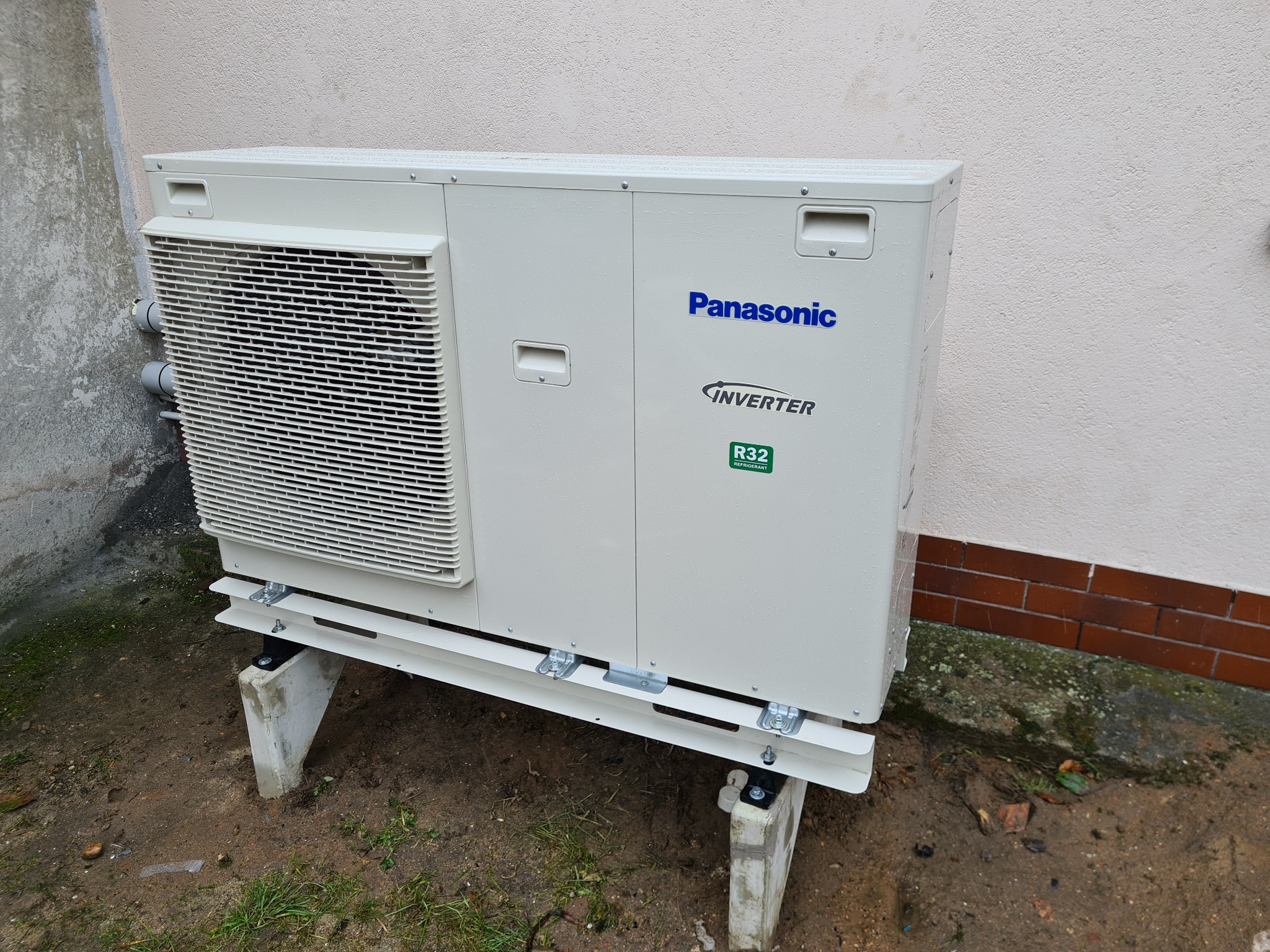 Zewnętrzna jednostka klimatyzacji Panasonic Inverter R32 zamontowana na betonowych podstawach przy ścianie budynku, widoczne ślady deszczu.