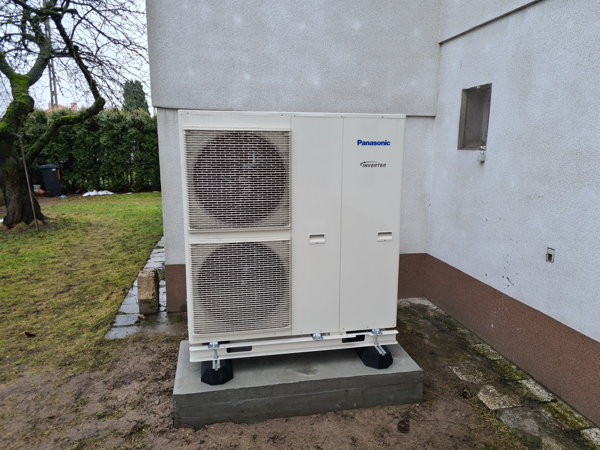 Zewnętrzna jednostka pompy ciepła Panasonic Inverter, zamontowana na betonowej podstawie przy ścianie budynku z widoczną kostką brukową i drzewem w tle.