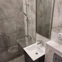 Nowoczesna łazienka z kabiną prysznicową typu walk-in, umywalką z szafką w kolorze grafitowym, lustrem w cienkiej ramie i toaletą z otwartą klapą, wykończona płytkami imitującymi marmur.