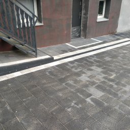 Seba - Ułożona szara kostka brukowa z odpływem liniowym wzdłuż budynku w Piekarach Śląskich, widoczne schody wejściowe z metalową barierką.