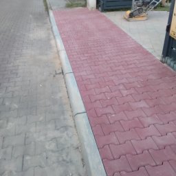 Seba - Świeżo ułożona ścieżka z czerwonej kostki brukowej o charakterystycznym wzorze, oddzielona szarym obrzeżem od starszej, szarej nawierzchni. W tle widoczna brama posesji i zagęszczarka do ubijania...
