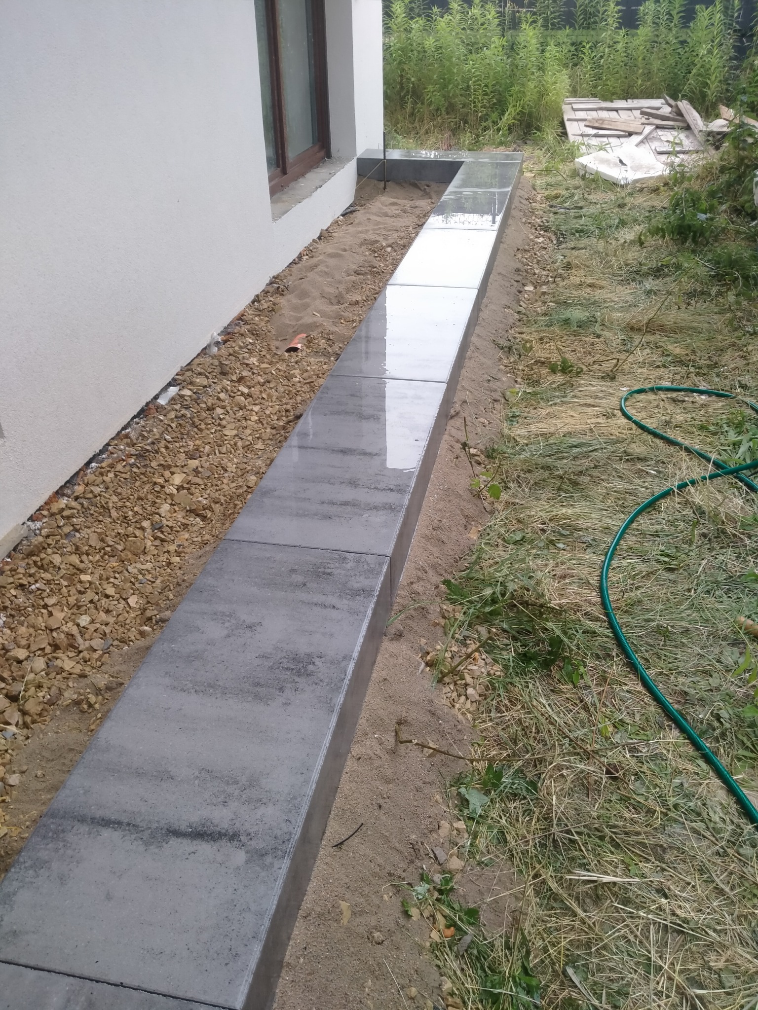 Świeżo ułożony, mokry chodnik z szarych, betonowych płyt obok ściany budynku, z widoczną warstwą piasku i kamieni po bokach oraz zielonym wężem ogrodowym w trawie.
