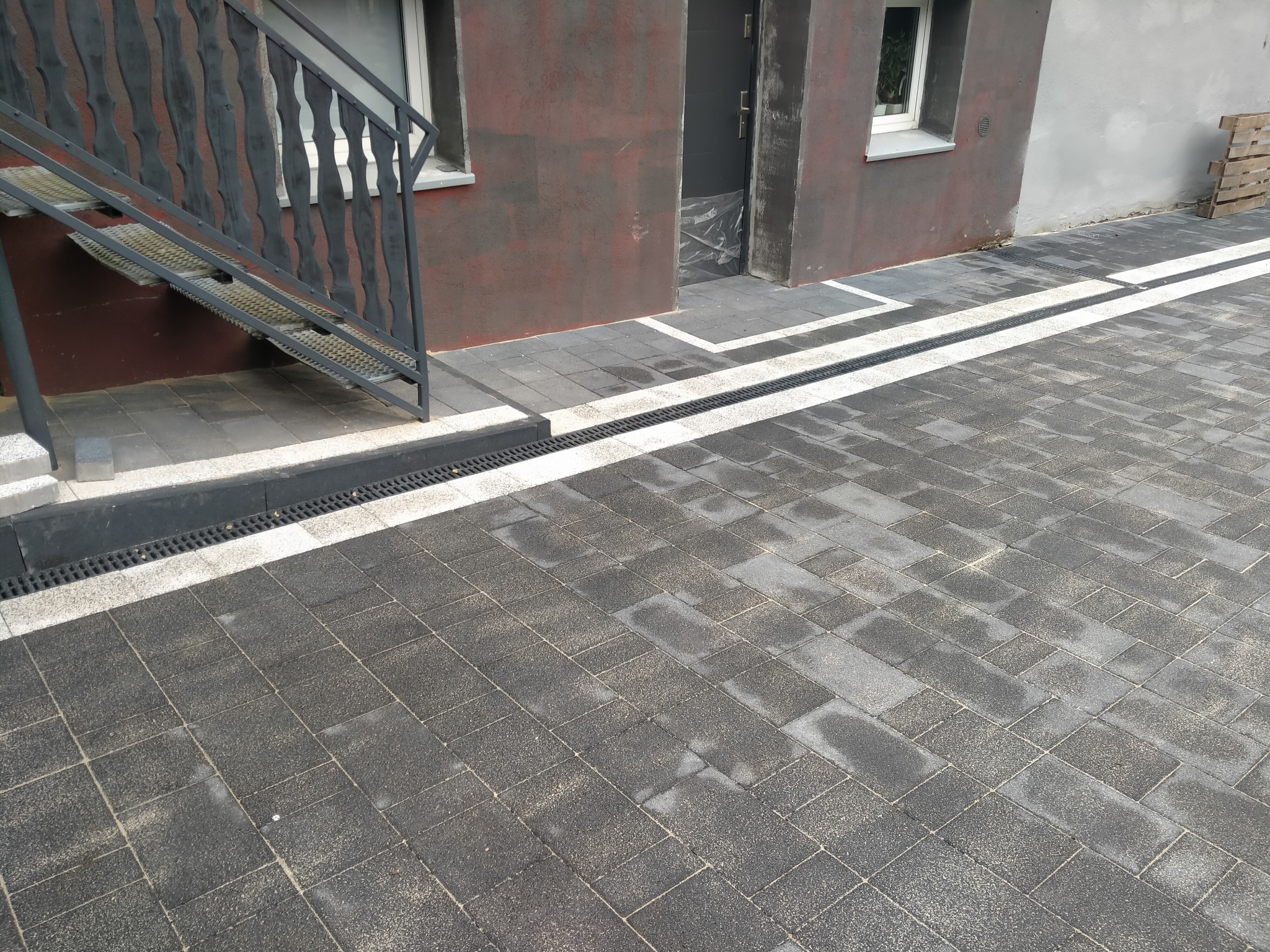 Ułożona szara kostka brukowa z odpływem liniowym wzdłuż budynku w Piekarach Śląskich, widoczne schody wejściowe z metalową barierką.