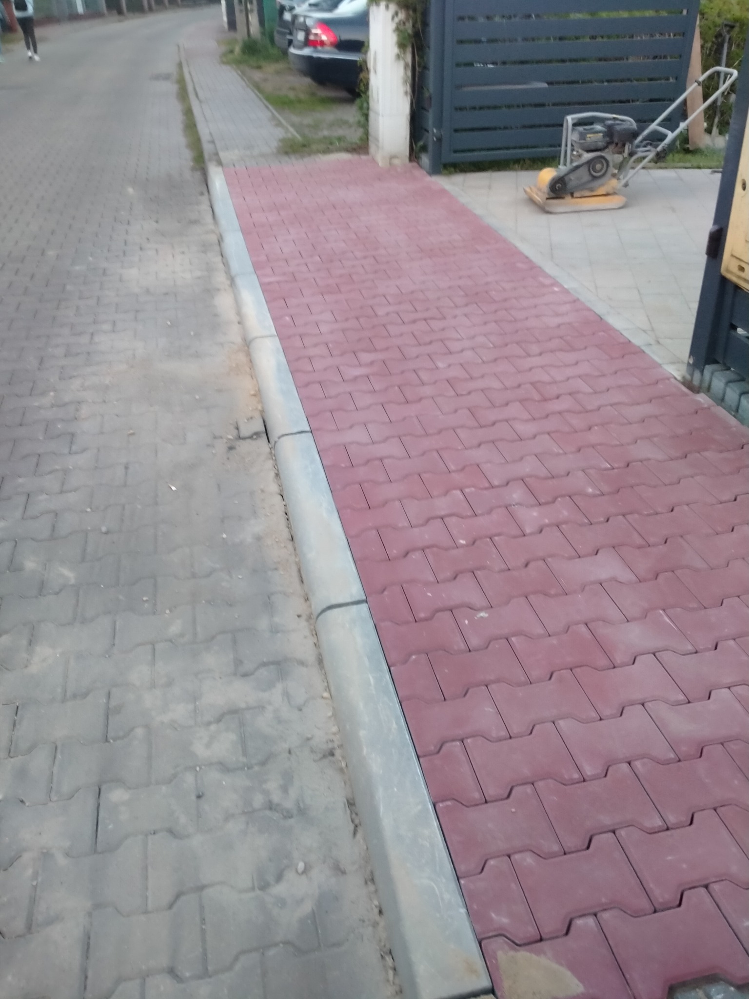 Świeżo ułożona ścieżka z czerwonej kostki brukowej o charakterystycznym wzorze, oddzielona szarym obrzeżem od starszej, szarej nawierzchni. W tle widoczna brama posesji i zagęszczarka do ubijania...