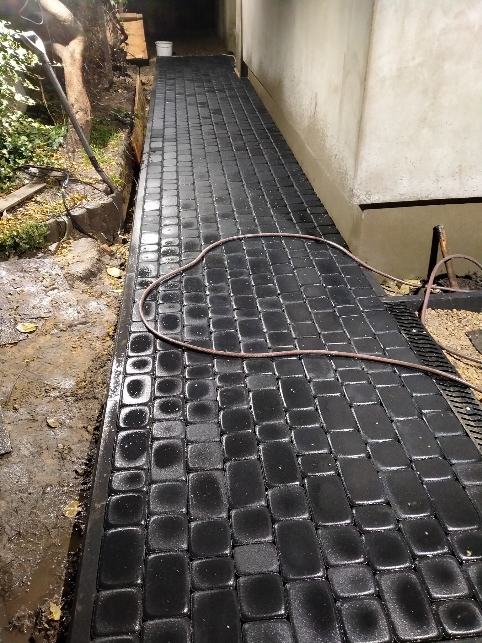 Ułożona czarna kostka brukowa z widocznym wężem ogrodowym na powierzchni, przylegająca do ściany budynku i ziemi z roślinnością.