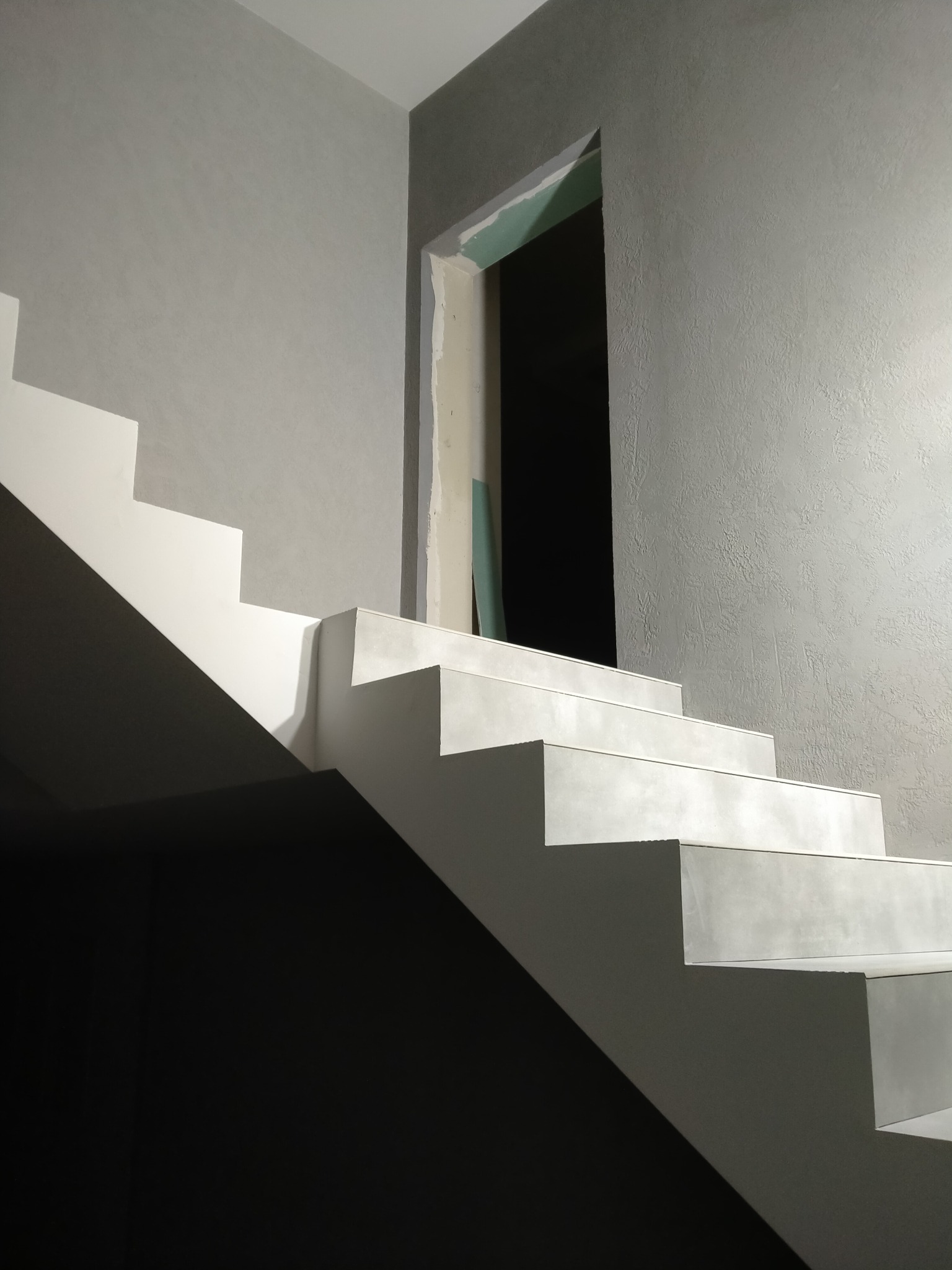 Nowoczesne, minimalistyczne schody wewnętrzne z białymi stopniami i szarą ścianą o wyraźnej fakturze, widoczne przygotowania do wykończenia ościeżnicy drzwiowej.