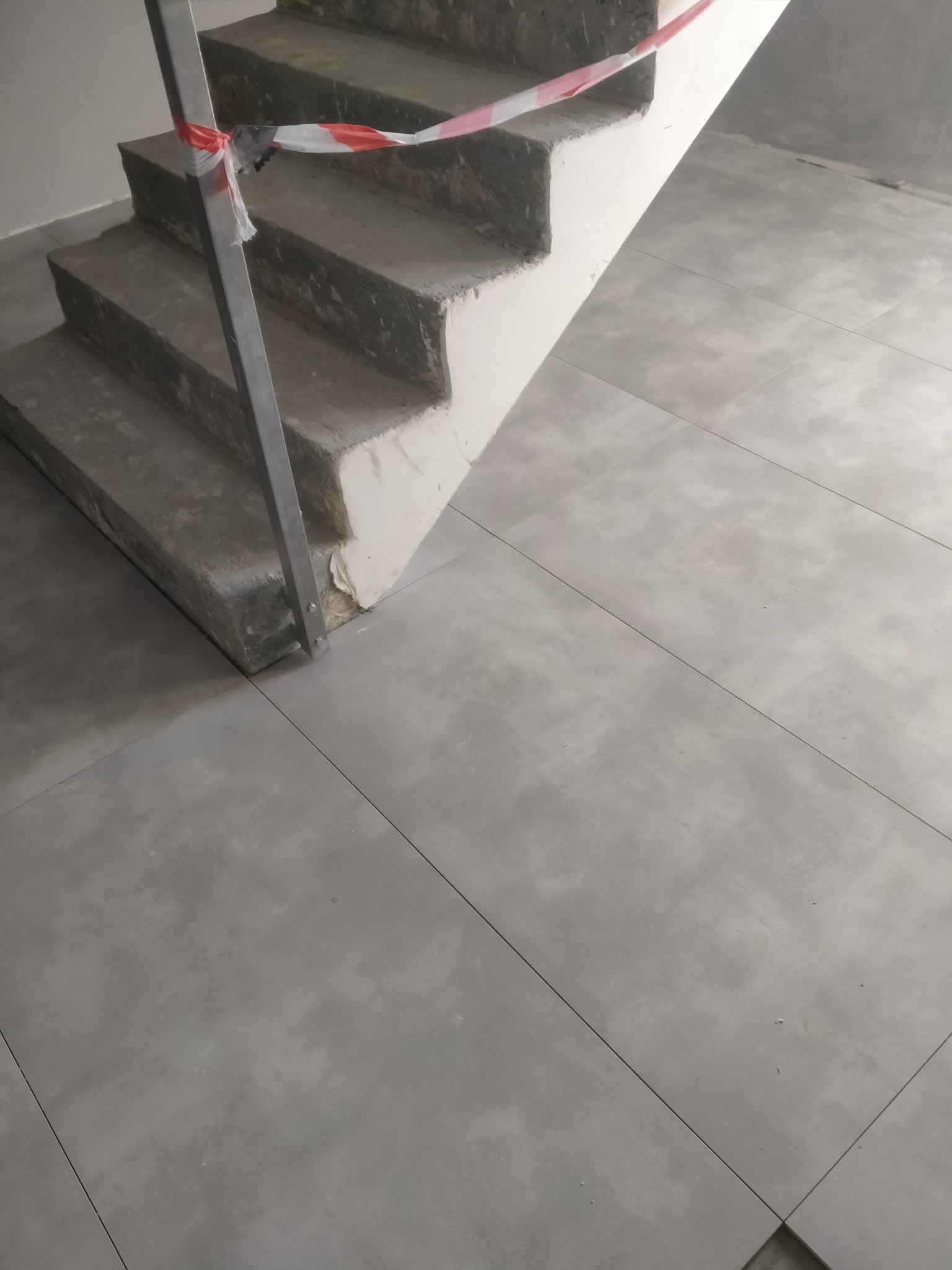 Surowe betonowe schody z metalową barierką i taśmą ostrzegawczą, kontrastujące z nowo ułożonymi, dużymi, szarymi płytkami podłogowymi.