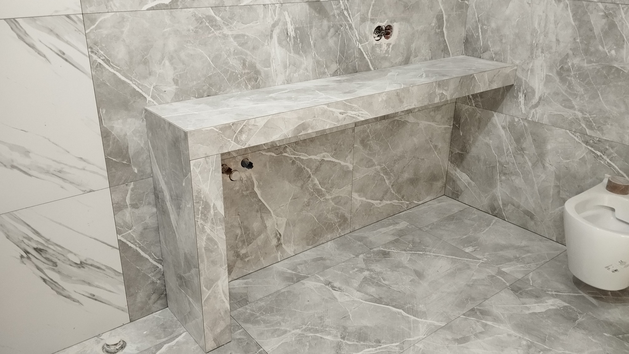 Nowoczesna łazienka z szarymi marmurkowymi płytkami, zabudowa umywalki z identycznych płytek, widoczne przyłącza hydrauliczne i elektryczne.