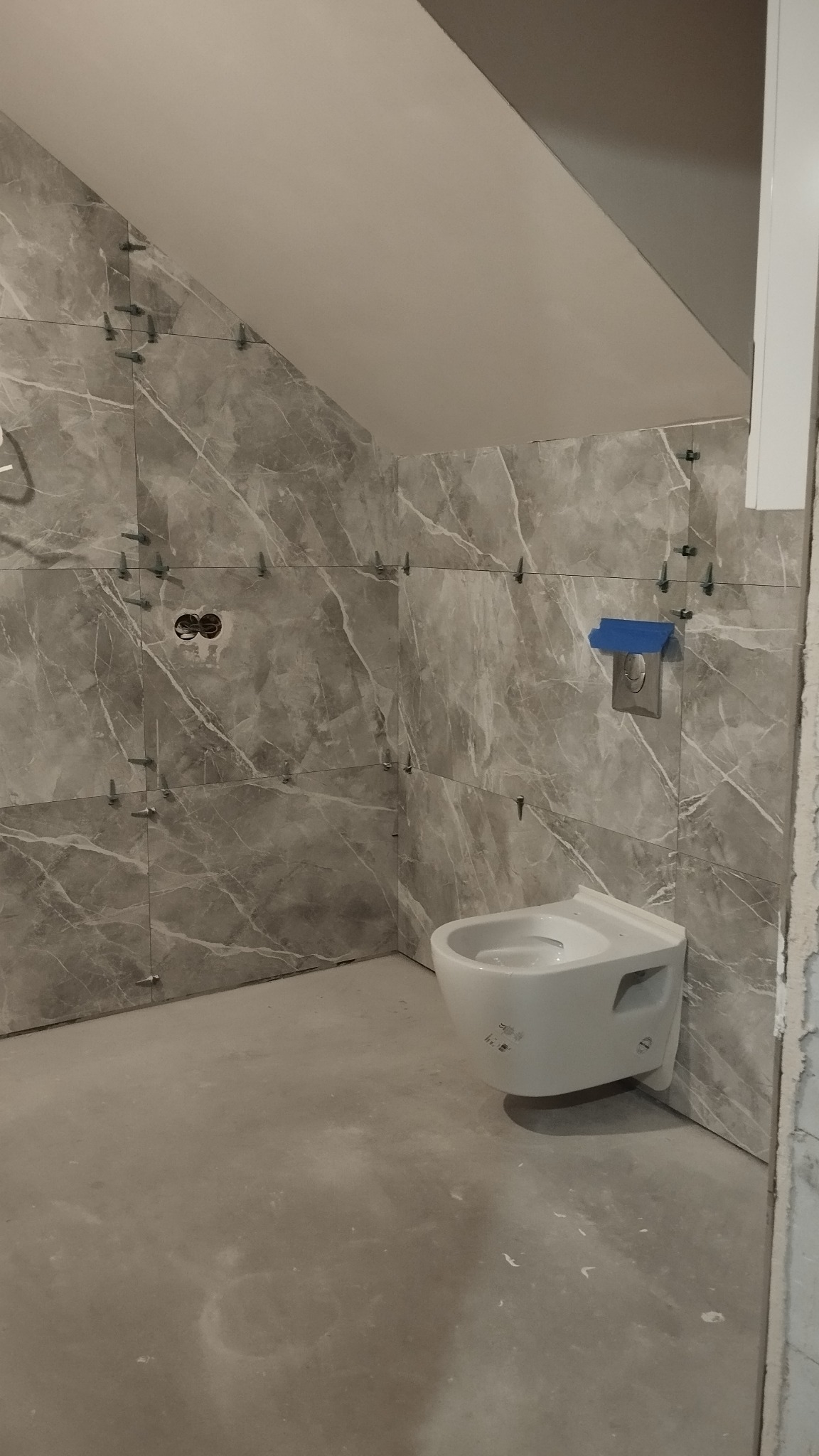 Łazienka w trakcie remontu: szare płytki imitujące marmur na ścianach z krzyżykami dystansowymi, zamontowana podwieszana toaleta, surowa podłoga przed położeniem płytek.